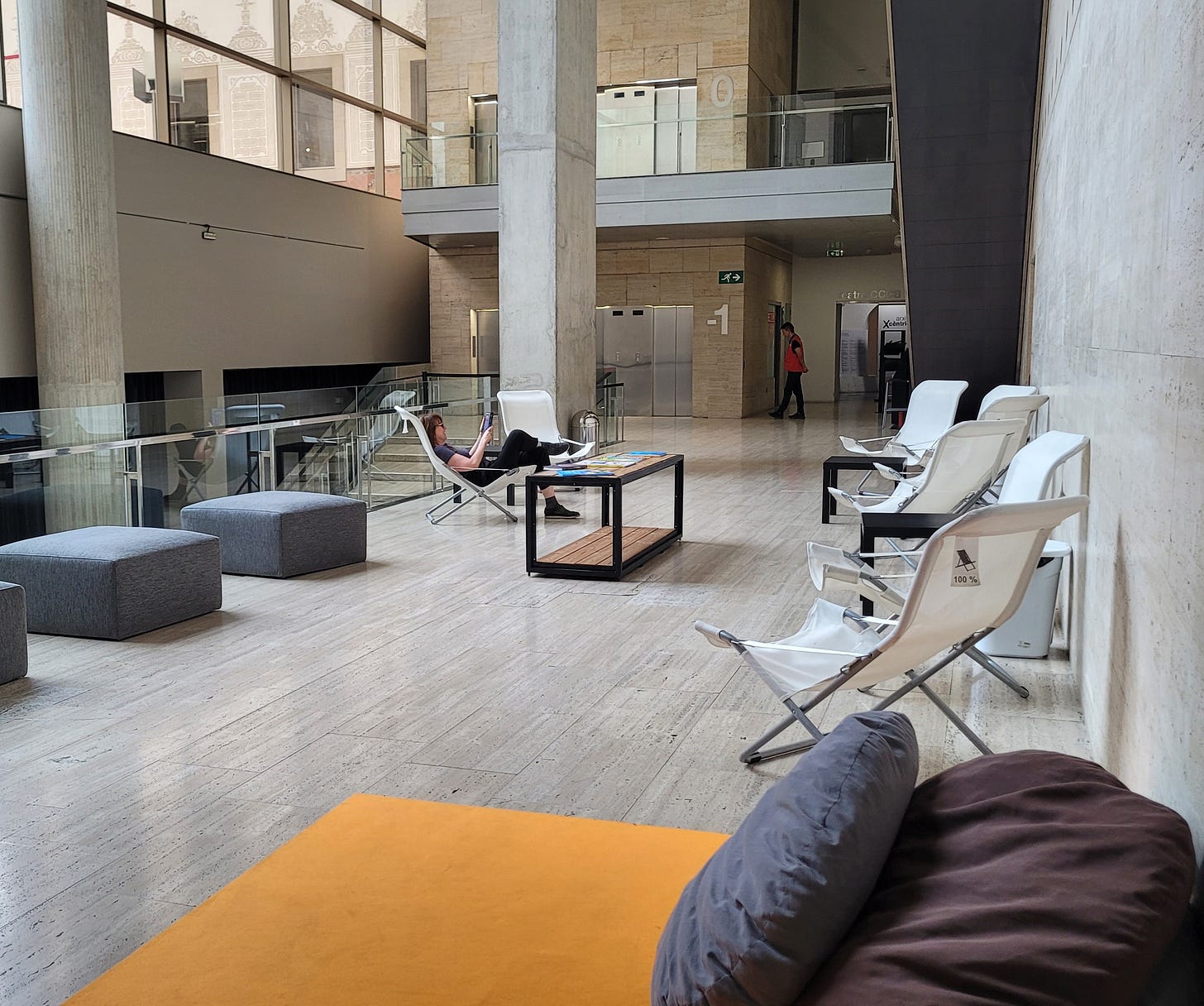 La sala all'ingresso del CCCB arredata con divanetti, sedie sdraio e un tavolino con giornali. Una persona sta leggendo un libro, accomodata su una sdraio.