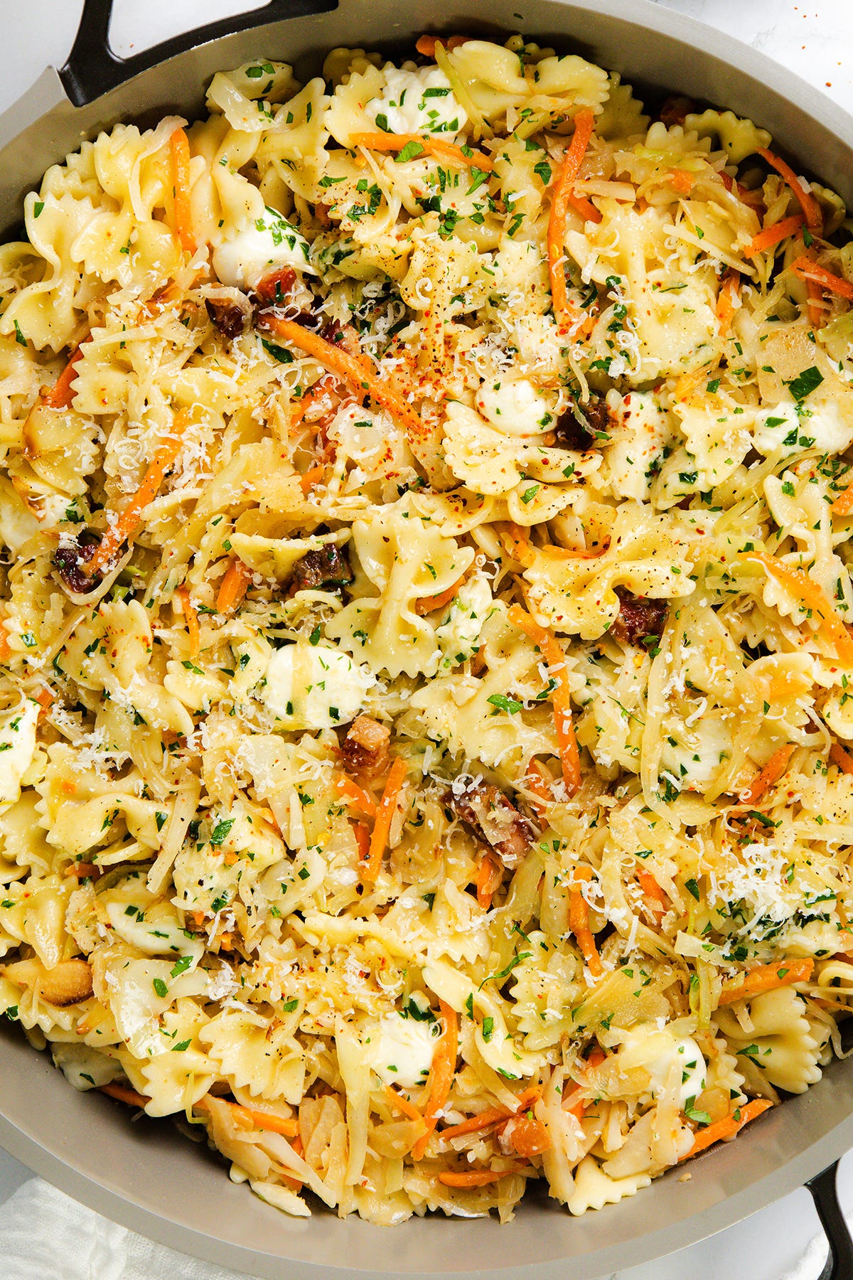 A skillet filled with bowtie pasta mixed with shredded carrots, cheese, herbs, and small pieces of sun-dried tomatoes, garnished with grated Parmesan and chopped parsley.