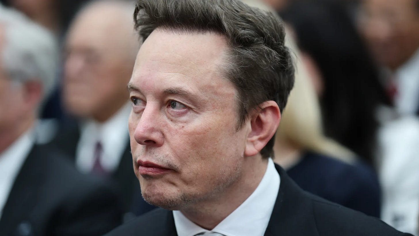 Elon Musk in a dark suit and white shirt looking to the side, with attendees seated behind him Elon Musk in a dark suit and white shirt looking to the side, with attendees seated behind him