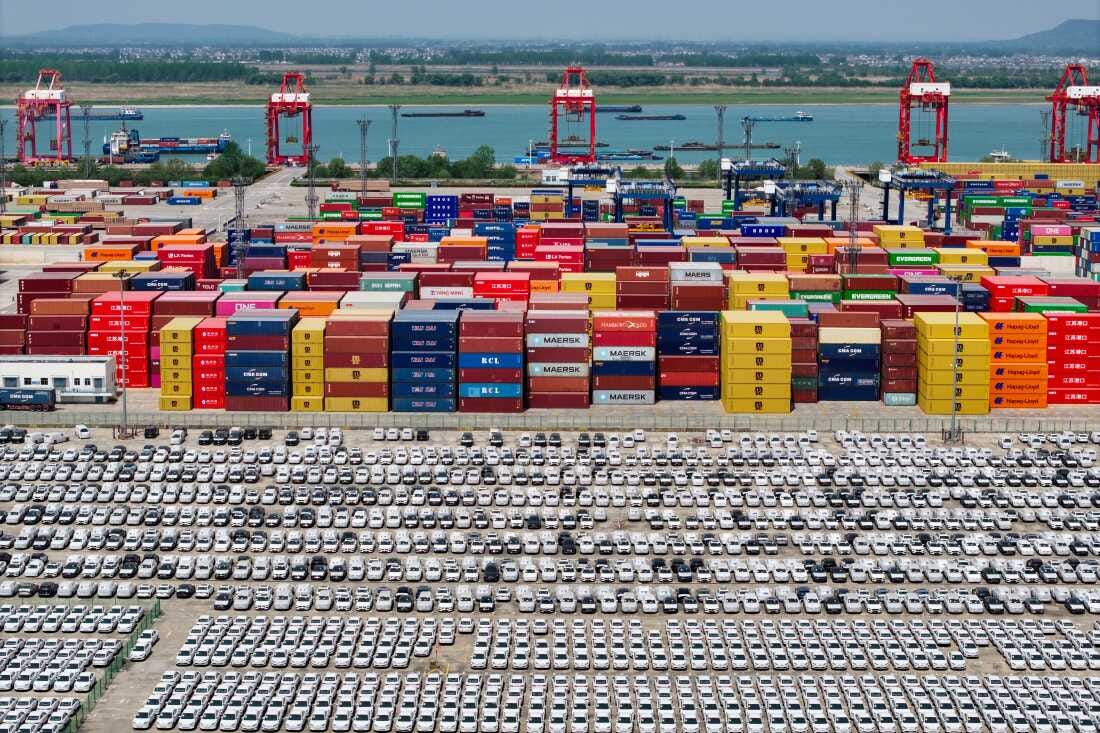 Chinese-made cars are lined up in rows at the port in Nanjing, China. Behind the cars are stacks of cargo shipping containers in a variety of colors — blue, red, yellow, brown, orange. Chinese-made cars are lined up in rows at the port in Nanjing, China. Behind the cars are stacks of cargo shipping containers in a variety of colors — blue, red, yellow, brown, orange.