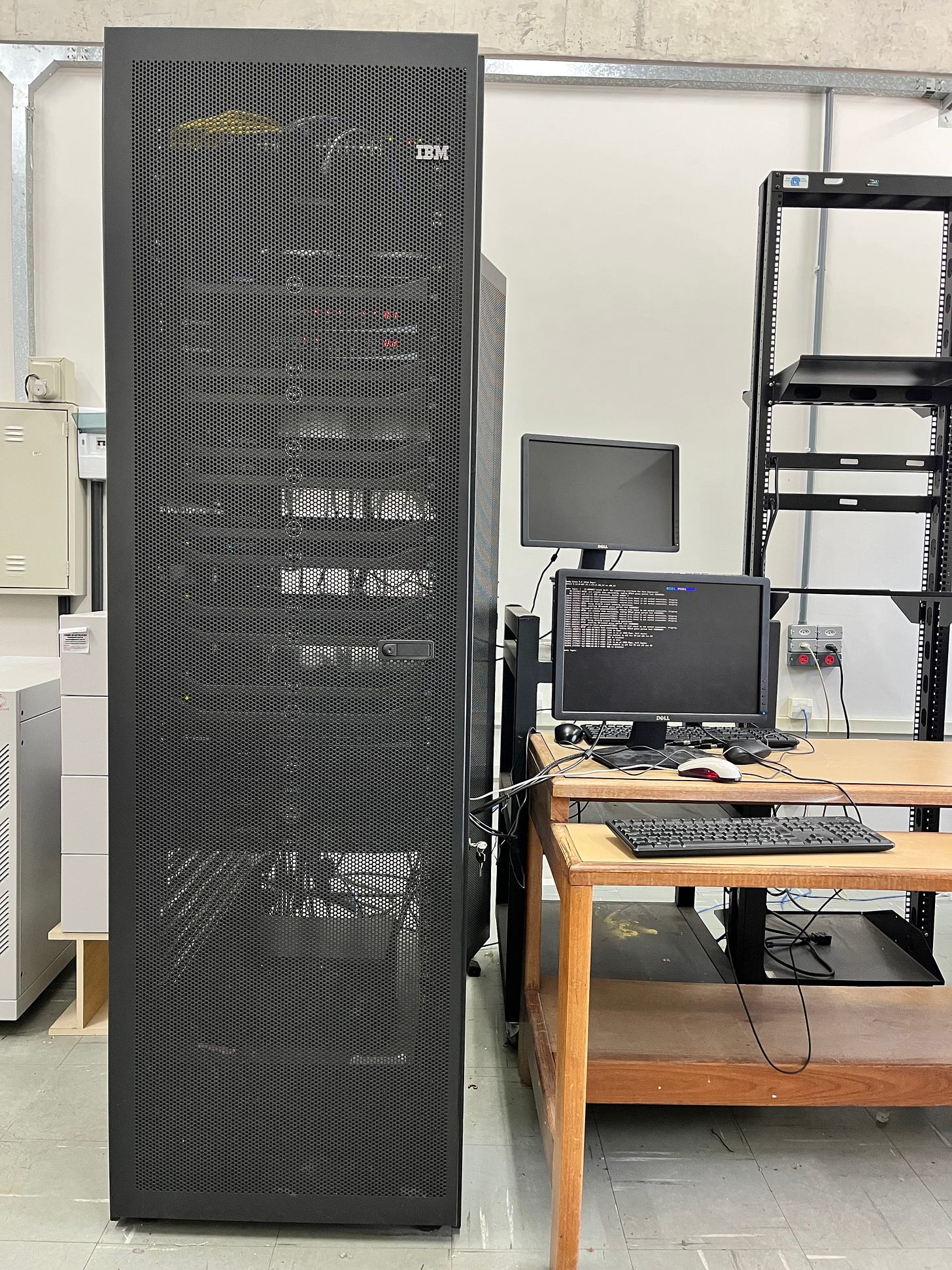 A tall, black mesh server rack filled with compute nodes stands in a university physics lab. To the right, a wooden desk holds a computer monitor displaying a Linux command-line terminal, a keyboard, and a mouse.