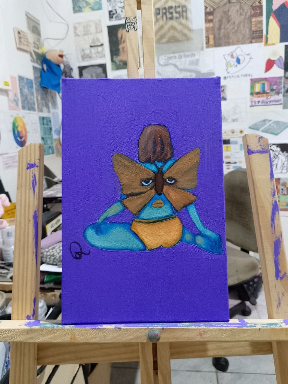 A imagem exibe uma fotografia de uma pequena pintura em um cavalete de madeira, em um ambiente desorganizado. A Pintura: A obra de arte, em primeiro plano na fotografia, apresenta uma figura feminina sentada no chão, de costas, com um corpo predominantemente azul. A figura tem uma grande borboleta marrom, com desenhos nas asas que se assemelham a olhos, pousada em suas costas. Abaixo da borboleta há uma pequena boca arqueada para baixo. O fundo da pintura é azul arrocheado. O Ambiente: A área ao redor do cavalete na fotografia é desorganizada, com papéis, pôsteres e outros itens visíveis ao fundo.