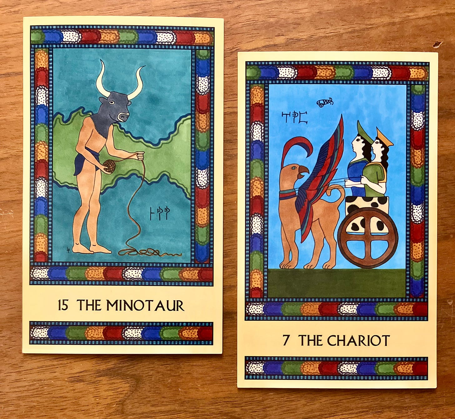 Two Minoan Tarot cards side by side on a brown wood surface. Both cards have intricate borders in deep multicolored tones. The Minotaur card, on the left, shows a bull-headed man wearing a loincloth. He is standing in shadowy darkness, facing the right, and unrolling a ball of thread, the free end of which pools at his feet. The Chariot card, on the right, shows a simple chariot covered in spotted cowhide, with a four-spoked wheel. The chariot is drawn by a griffin with brightly colored wings. In the chariot ride two robed female figures wearing hats. Both cards have small Linear B inscriptions near the main figures.