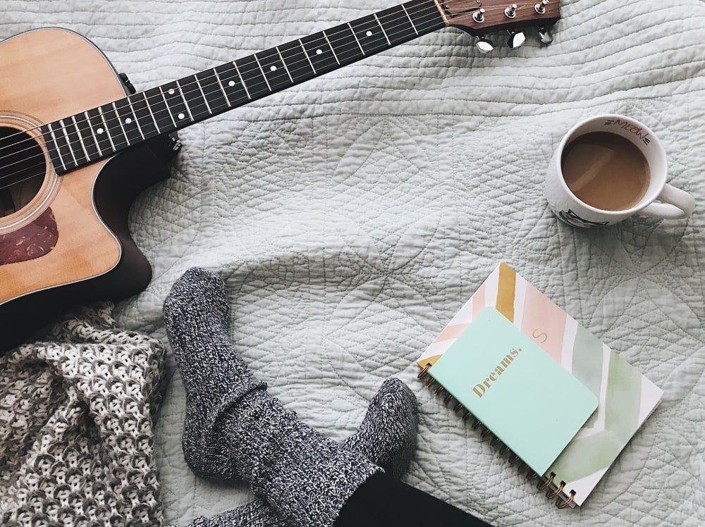 Persona con calcetines cómodos junto a guitarra acústica, cuaderno con "dreams" en la portada y una taza de café, todo ello sobre una colcha de cama Persona con calcetines cómodos junto a guitarra acústica, cuaderno con "dreams" en la portada y una taza de café, todo ello sobre una colcha de cama