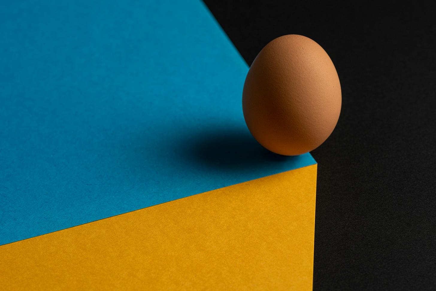 Un uovo in bilico sullo spigolo di una superficie azzurra, con la parete verticale visibile color giallo e lo sfondo nero. Un uovo in bilico sullo spigolo di una superficie azzurra, con la parete verticale visibile color giallo e lo sfondo nero.