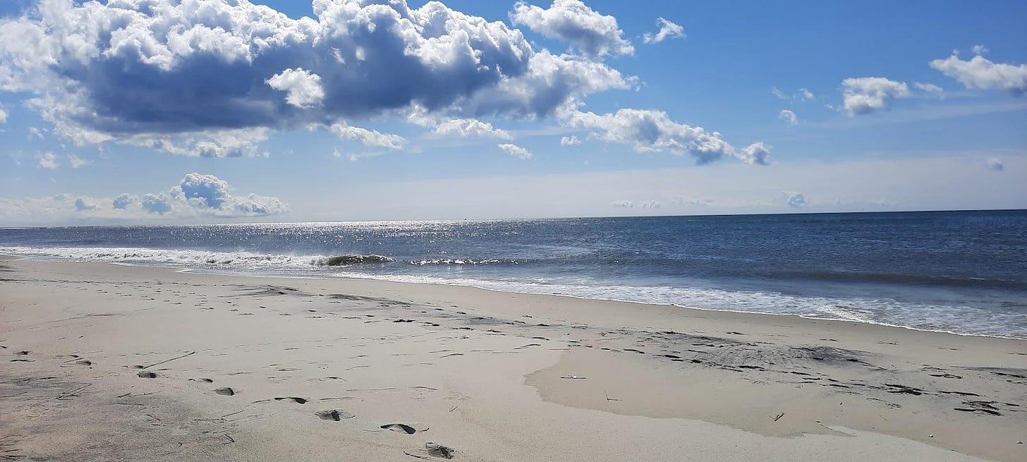 A sandy beach and bright blue sea, reflecting sunlight to the top left. There are footsteps walking away from the camera to the left.