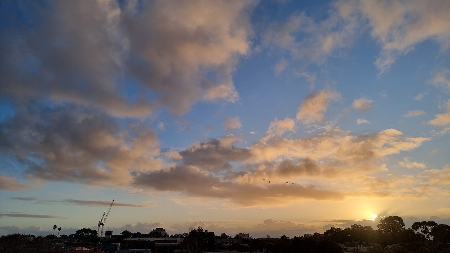 A photo of a sunset with fluffy clouds of gray, white and yellow against a gorgeous blue sky. Some birds are silhouetted against some clouds.