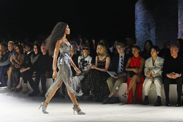 A model in a silver gown is shown in profile as she strides along a runway in front of a line of audience members.