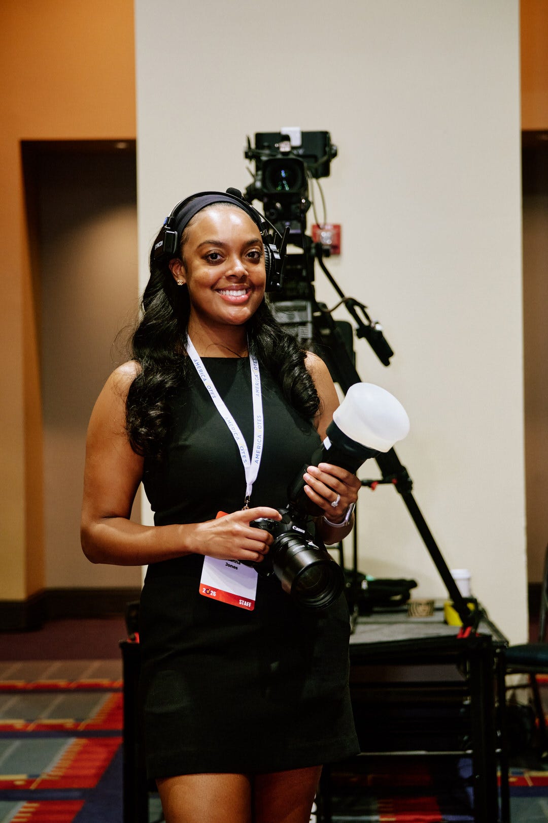 Photographer Jones from Annan Productions in all black with headphones around her neck, holding a Canon camera body and white flash diffuser and smiling, a large broadcast camera rig behind her
