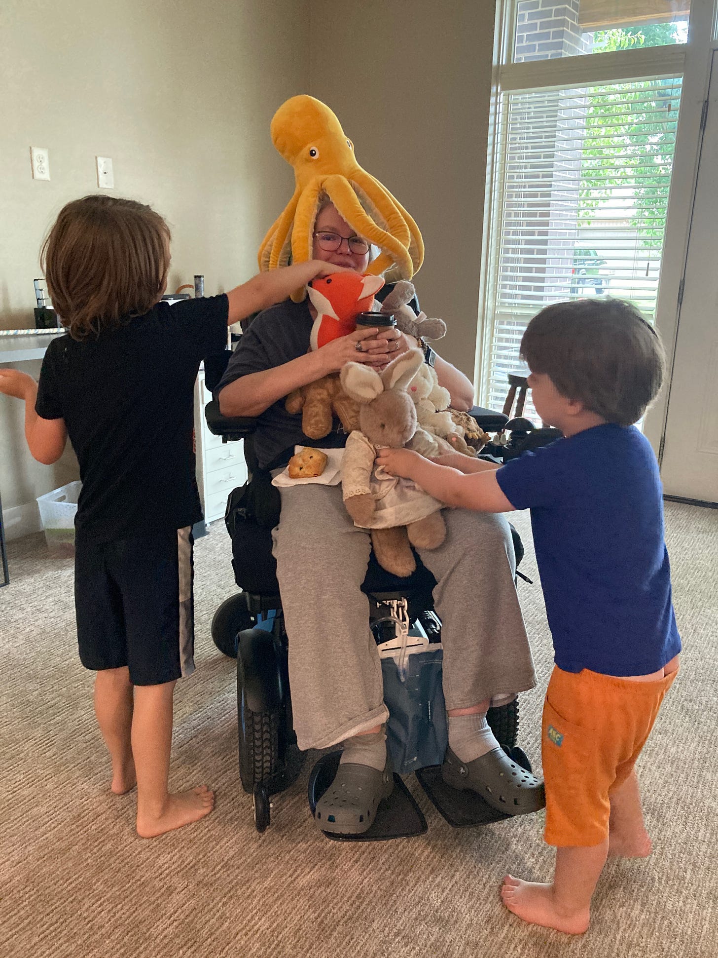 Debbie is in her living room and two small children are covering her with stuffed animals while she thinks of drinking her coffee and eating a scone that is resting on her knee