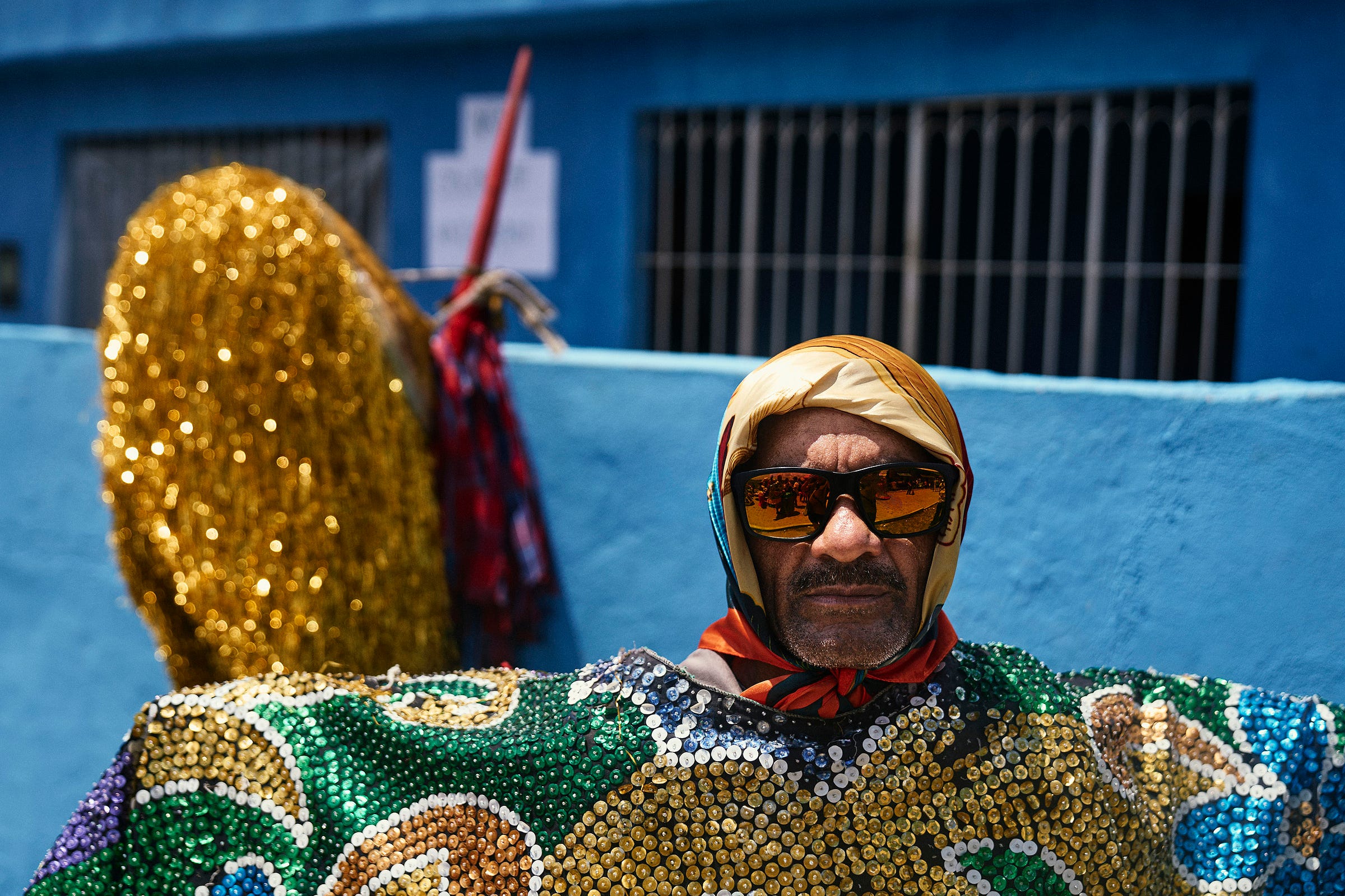 A person wearing reflective sunglasses, a colorful sequined outfit, and a gold headscarf stands in front of a bright blue wall with barred windows and a large golden decorative object behind them.