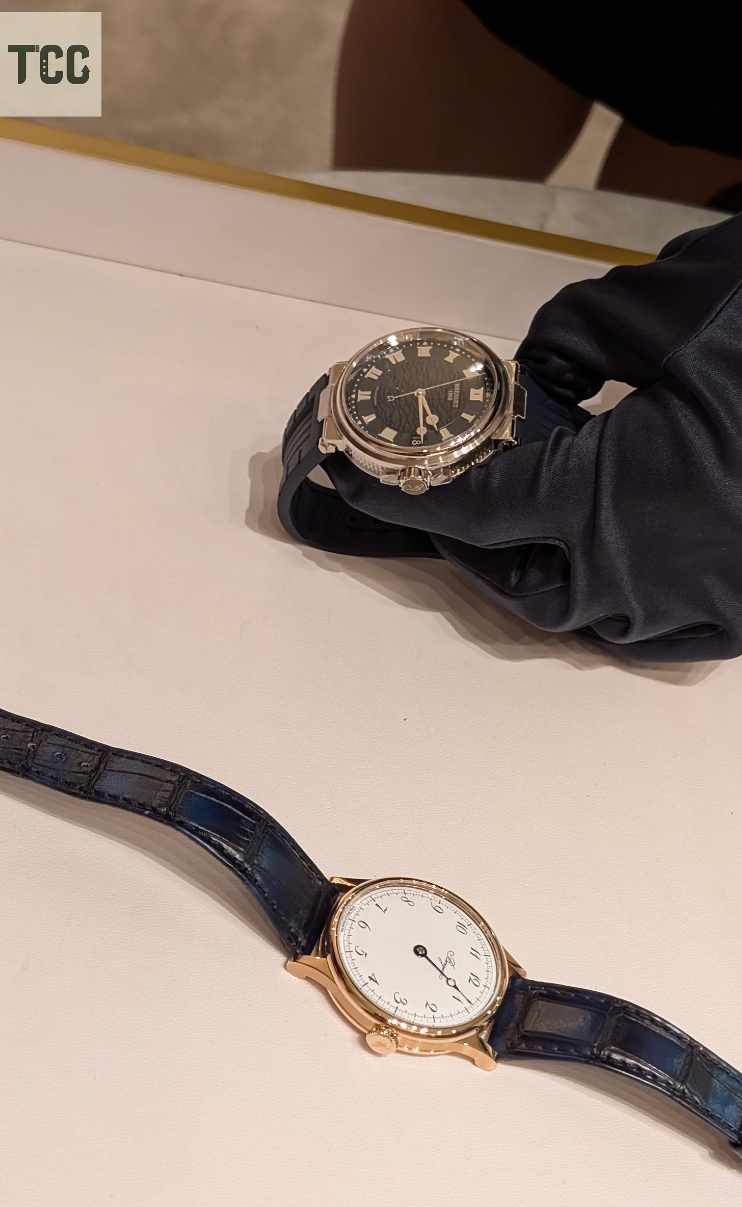 Close-up of two Breguet watches on a display counter: a silver Breguet Marine with a textured dark dial held by a gloved hand, and a rose gold Breguet Souscription watch with a white dial and a single hour hand on a blue alligator strap.