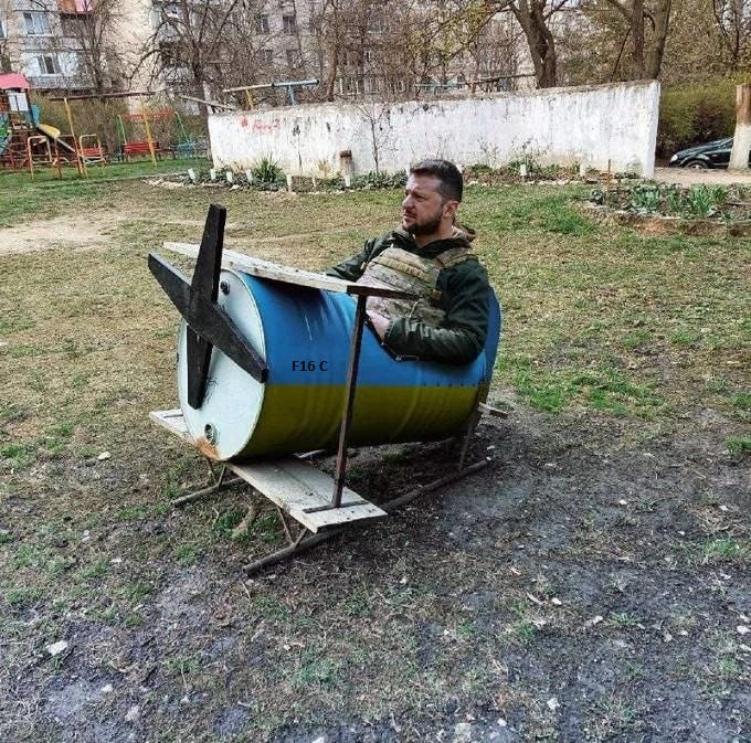 A man sitting inside a makeshift structure resembling an airplane, constructed from a blue and yellow barrel with a propeller attached to the front. He is wearing a green jacket and tactical vest, positioned on a grassy field near a playground and residential buildings. The setup humorously mimics a surveillance drone, identified as the Ghost of Kiev in the context.