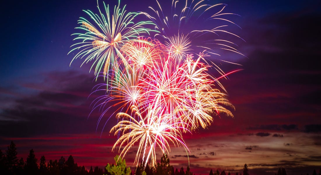 Green, purple, and orange fireworks in the night sky, with a purple and pink sunset