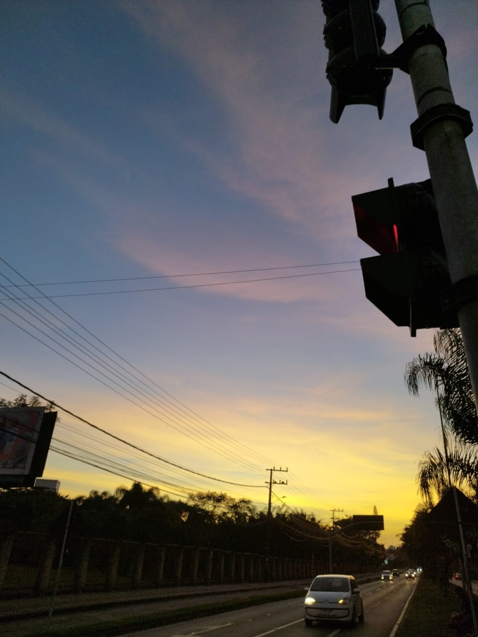 Foto de um pôr do sol azul com traços de amarelo. Em primeiro plano, um semáforo e um carro na rua.