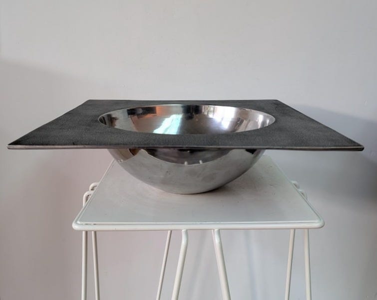 May include: A bowl featuring a polished, reflective stainless steel basin integrated into a flat, square, dark gray matte metal frame. The bowl has a smooth, curved interior and exterior surface, contrasting with the textured, flat square border.