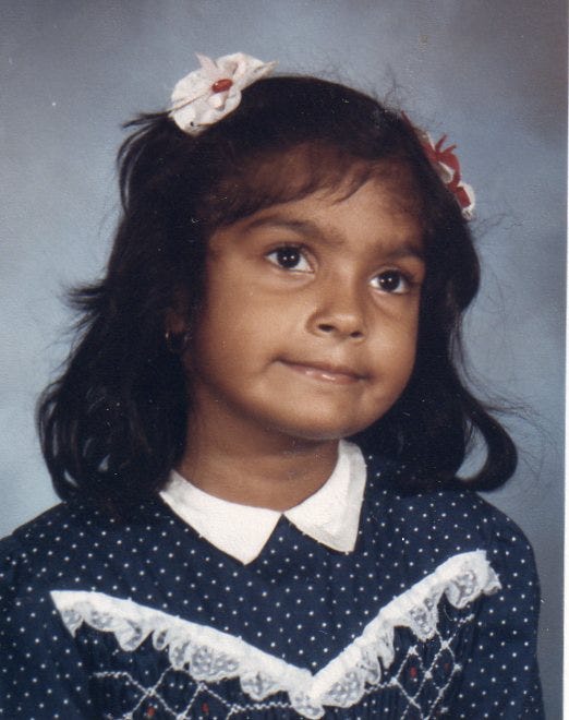 A school photo of Nisha as a young girl, probably in 2nd grade. She has a navy blue dress with white dots and lace. She smiles with her lips closed and head tilted.