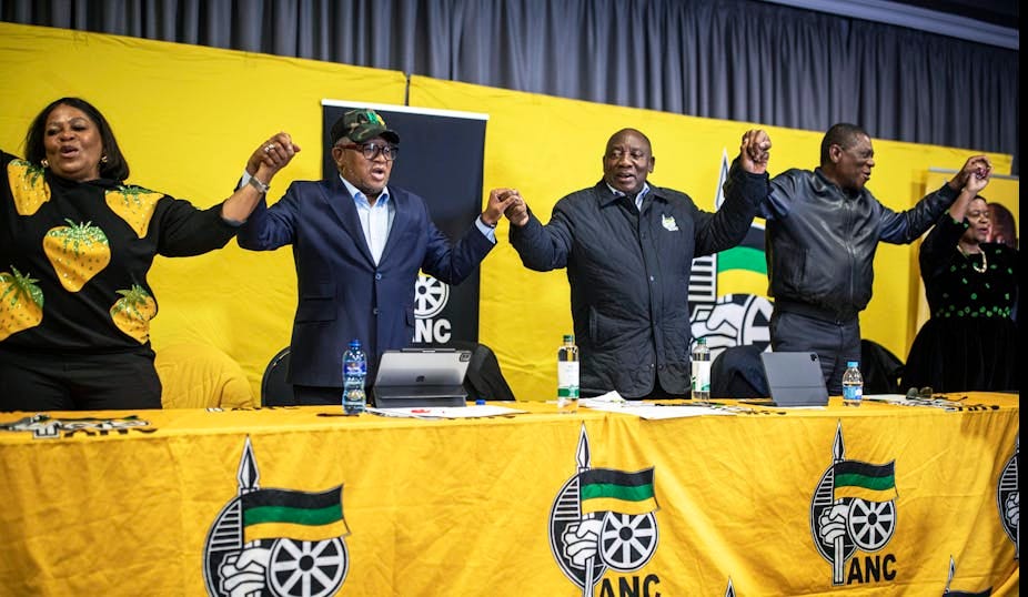 Three men and two women stand at a long table draped in yellow fabric with the ANC insignia. They are holding hands and appear to be singing or chanting. Three men and two women stand at a long table draped in yellow fabric with the ANC insignia. They are holding hands and appear to be singing or chanting.