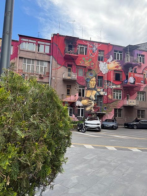 Scenes from around Armenia: bronze deer sculptures near the Cascade complex in Yerevan, a stone monastery nestled against forested mountains, a glass of amber brandy resting on an aging barrel, a hand holding a wine glass and folded lavash wrap at an outdoor food stand, bunches of grapes drying for raisining, and a colorful mural covering an apartment building facade in Yerevan