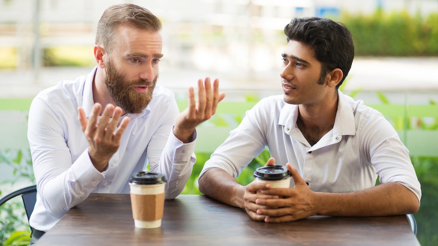 Two men sit at an outdoor table enjoying coffee while they talk. Two men sit at an outdoor table enjoying coffee while they talk.