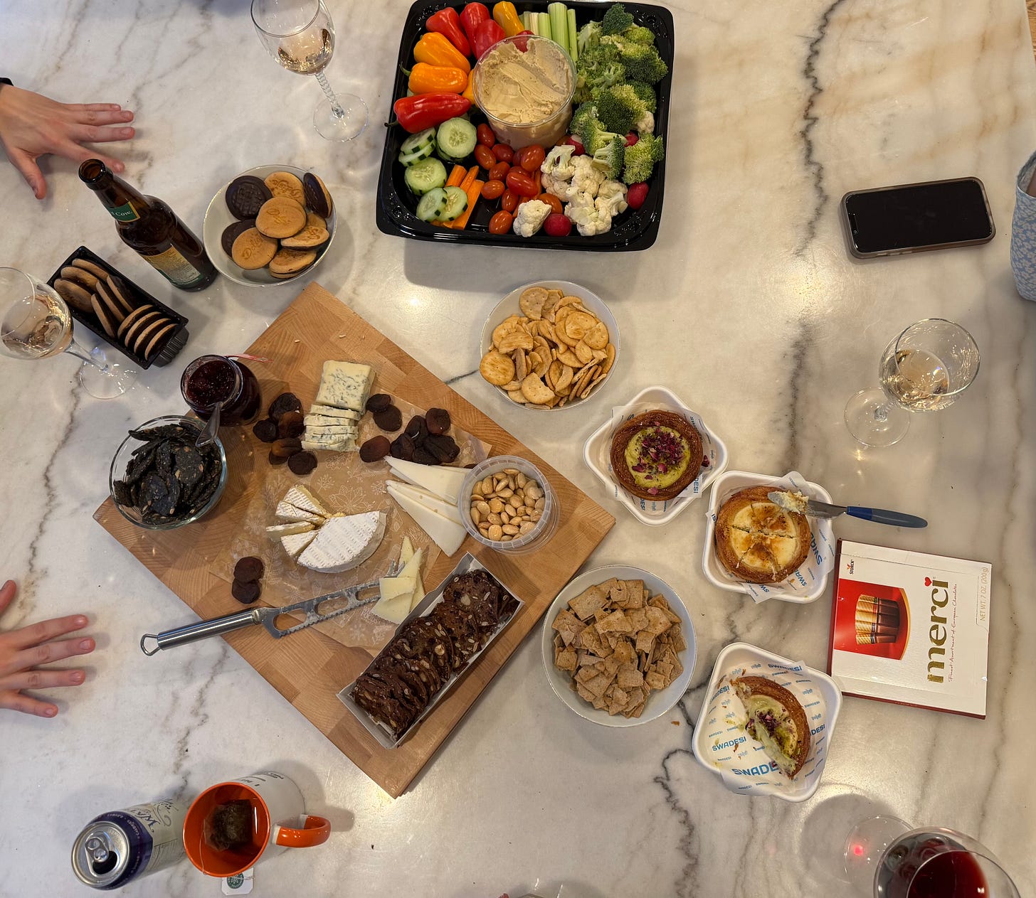Holiday grazing table with vegetables, cheeses, crackers, sweets, and drinks spread across a countertop, surrounded by people’s hands.