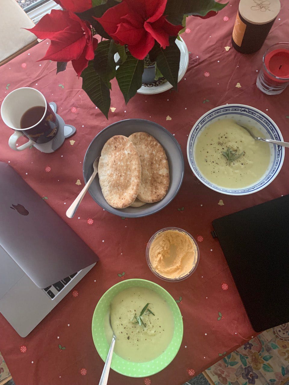 A top-down photo of a dining table covered by a red Christmas tablecloth, bowls of soup, mugs of tea, a bowl of pita bread, and candles