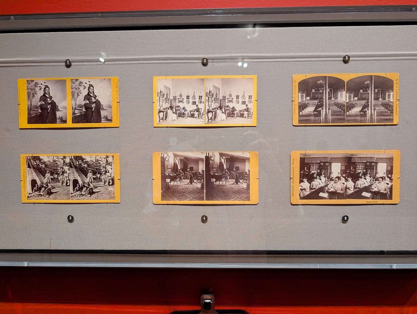 Display case with six stereoscopic photo cards from the 1860s to 1890s, including portraits of Indigenous people, interior scenes, classrooms, and parlor exhibitions, all mounted on yellow-bordered cards.