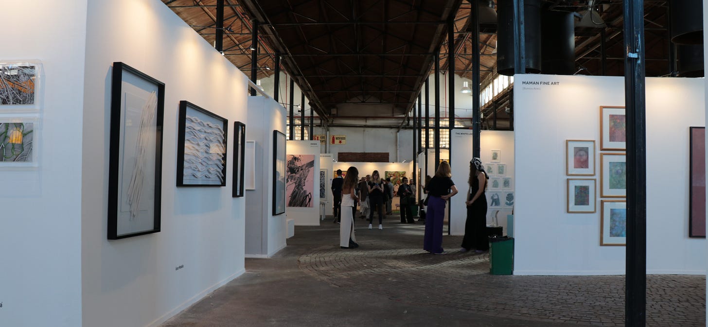 Interior de la Feria MAPA en el predio de La Rural durante su edición del año 2024. La imagen muestra un amplio espacio con techos altos y estructura de acero. Varias personas, predominantemente mujeres, caminan entre las paredes blancas que exhiben diversos cuadros y obras de arte. La iluminación suave complementa la atmósfera serena del lugar.