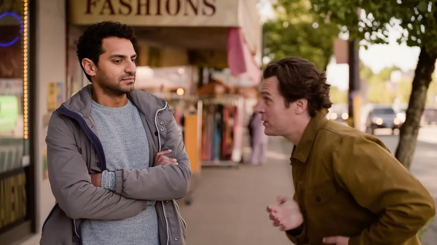 Naveen crosses his arms and looks unimpressed as Jay animatedly sings Bollywood show tunes  to him on a city sidewalk outside a fashion store.