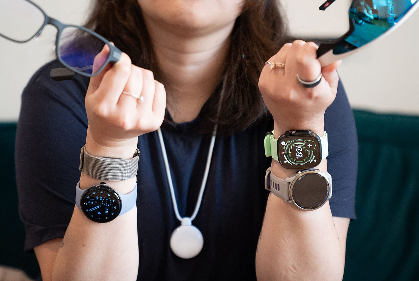 Close up of Senior reviewer Victoria Song holding two pairs of smart glasses, wearing four wrist-based wearables, a smart ring, and the Friend AI necklace.