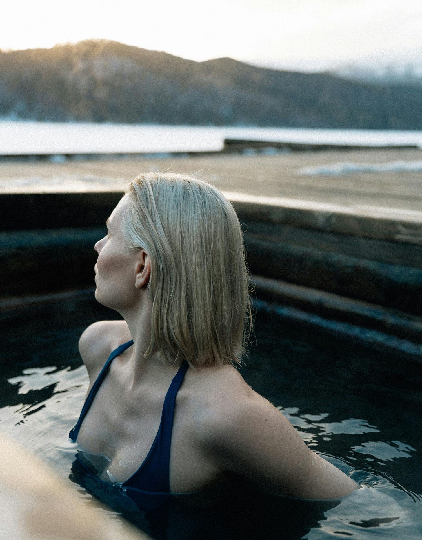 A woman with light blonde hair, wearing a dark swimsuit, sits in an outdoor cold plunge pool, gazing away from the camera. Snowy mountains are visible in the background, suggesting a chilly, serene setting. A woman with light blonde hair, wearing a dark swimsuit, sits in an outdoor cold plunge pool, gazing away from the camera. Snowy mountains are visible in the background, suggesting a chilly, serene setting.