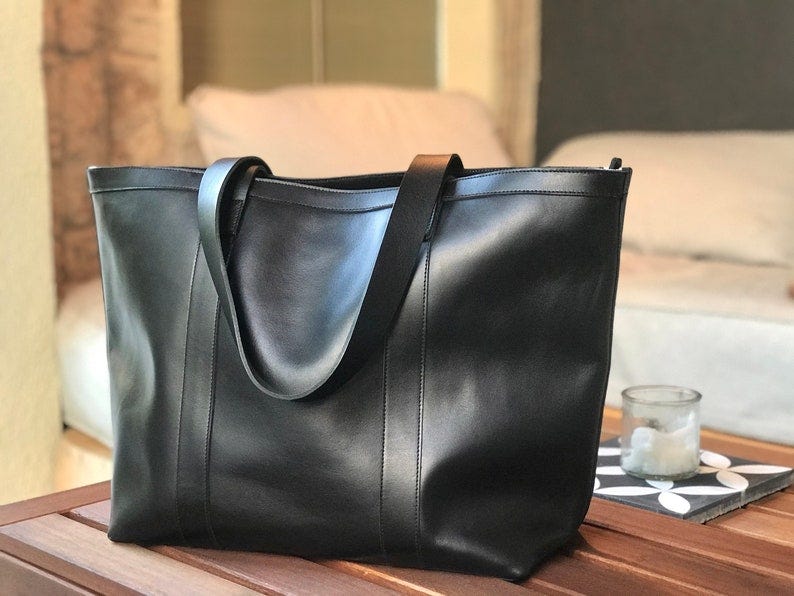 May include: A black leather tote bag with two wide straps. The bag is sitting on a wooden surface.