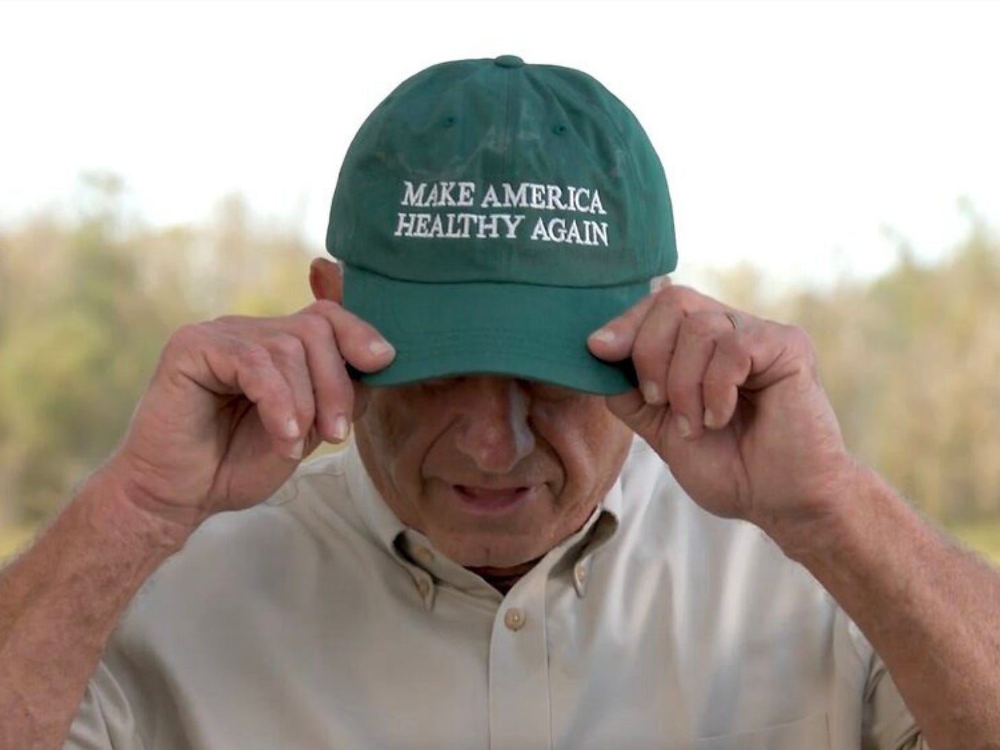 RFK Jr. pulling a green baseball cap embroidered with 'Make America Healthy Again' down over his eyes outdoors.