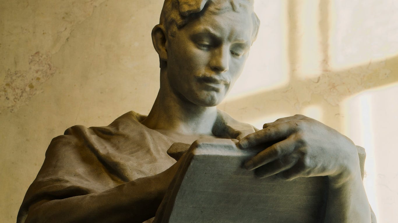 A moustachioed statue writes on a stone tablet, or maybe paper - but it's stone.