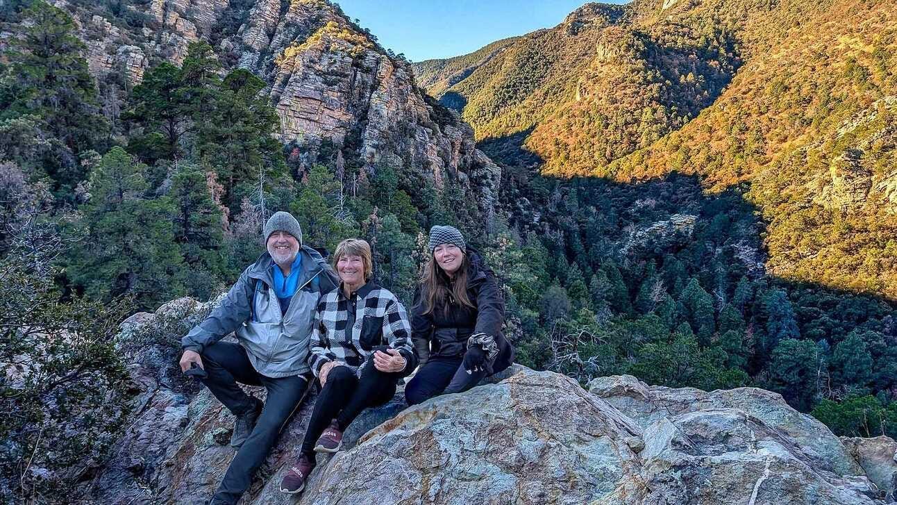 3 people sitting on a mountainous rock overlooking a valley of pine trees as the first rays of sunlight hit a neighboring mountain;
