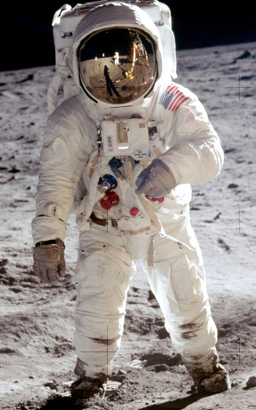 A photograph of astronaut Buzz Aldrin standing on the Moon during the Apollo 11 mission in 1969. He is wearing a bulky white spacesuit with a large helmet, and his visor reflects the lunar module and fellow astronaut Neil Armstrong. The Moon’s surface appears rocky and desolate beneath him.