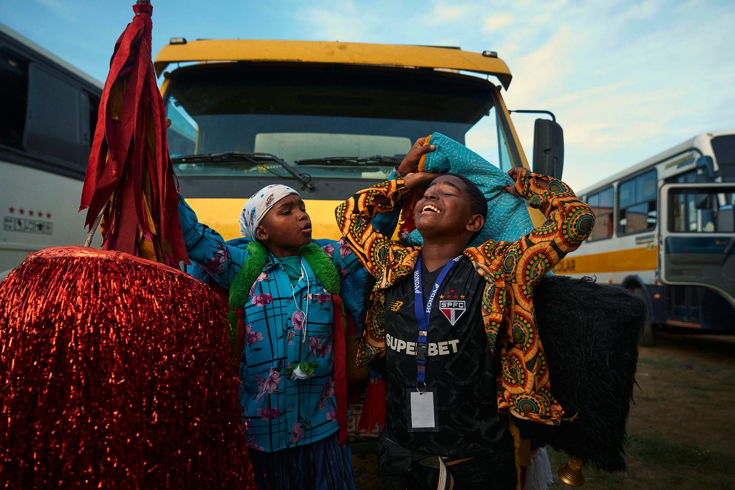 Two children stand in front of a yellow bus, smiling joyfully. One wears a colorful shirt and headscarf, while the other, in a patterned jersey, holds festive costumes in red and black. Buses are parked in the background.