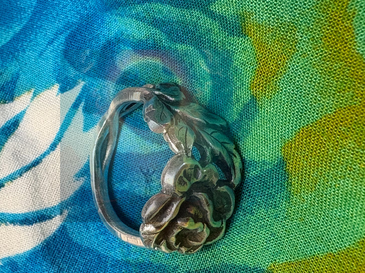 A silver ring, with a twisted, leaf-like design and small rose detail sits on a blue, green, and yellow patterned fabric. 