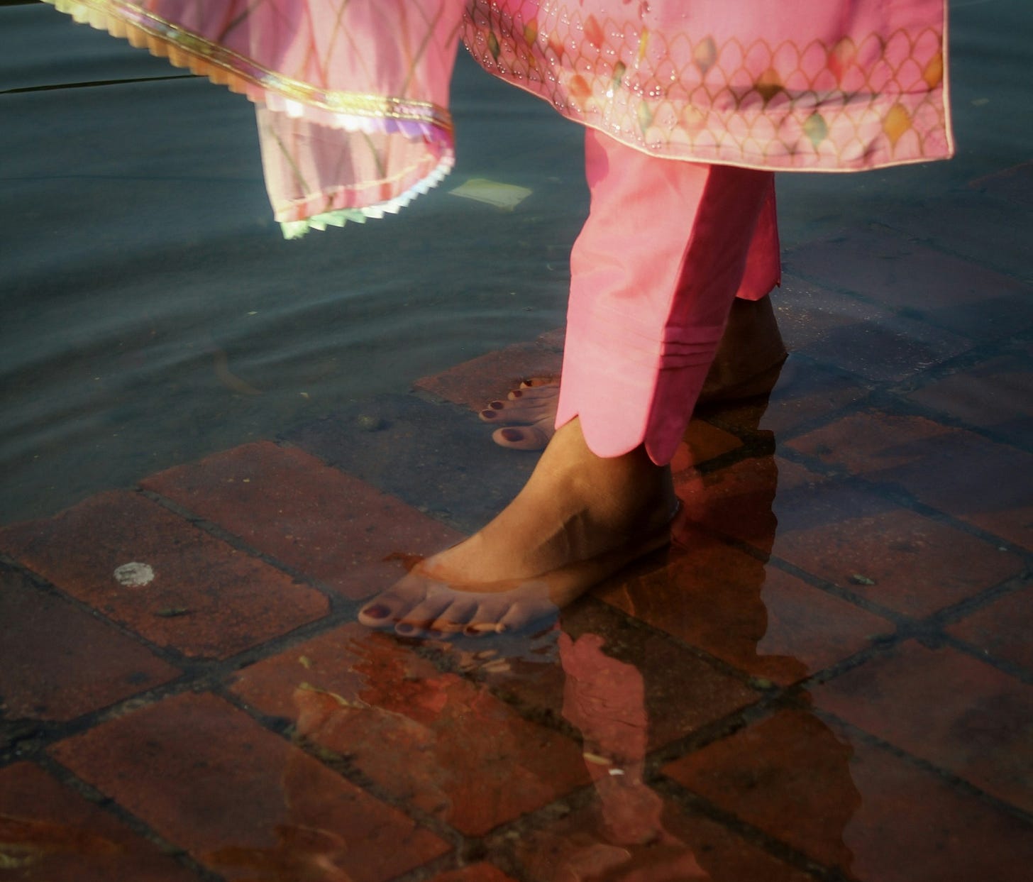 an indian woman walks on bricks submerged in water as her pink salwaar kameeze floats above her ankles