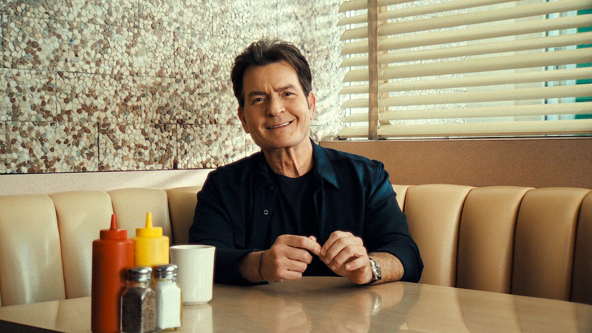 Charlie Sheen sits in a retro-style diner booth, smiling slightly with his hands folded on the table beside condiments and a coffee cup. Charlie Sheen sits in a retro-style diner booth, smiling slightly with his hands folded on the table beside condiments and a coffee cup.