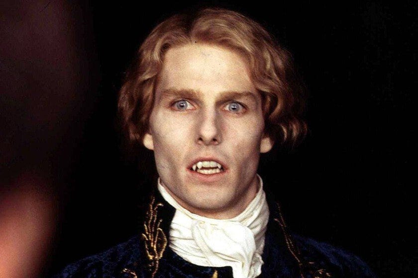 O ator Tom Cruise em close, com lentes azuis bem claras nos olhos, a boca entreaberta deixando ver os caninos afiados de vampiro e o cabelo loiro ondulado amarrado para trás. Vemos alguma espécie de casaca de veludo azul com bordados dourados, por cima de uma gola branca.