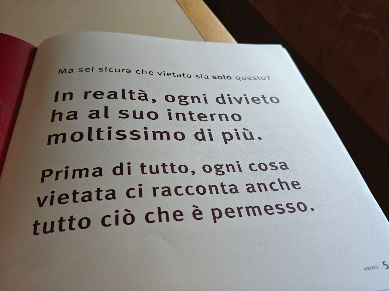 Una pagina aperta di un libro, la pagina 5 delle Scatoline di effequ intitolata "Vietato", che mostra un testo stampato su carta bianca. Il testo è in italiano: "Ma sei sicurə che vietato sia solo questo? In realtà, ogni divieto ha al suo interno moltissimo di più. Prima di tutto, ogni cosa vietata ci racconta anche tutto ciò che è permesso."  Si nota l'ombra di un tavolo o di una superficie di legno nell'angolo in alto a destra e parte della copertina rossa del libro sul lato sinistro.