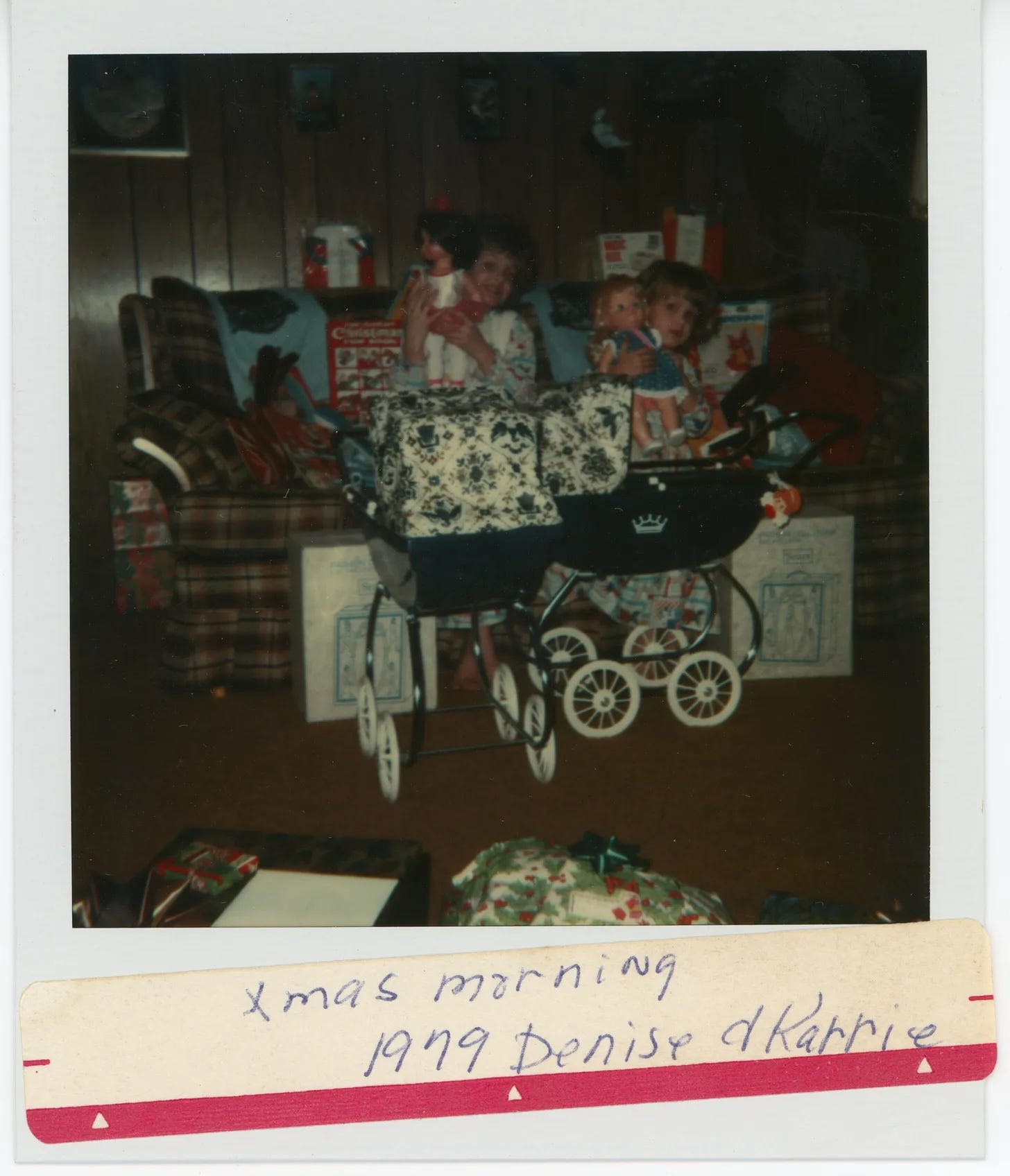 Karrie and her older sister in 1979 posing in front of two blue strollers holding up their dolls; the sister is excited and smiling and showing off her doll, while Karrie looks timid and unhappy.