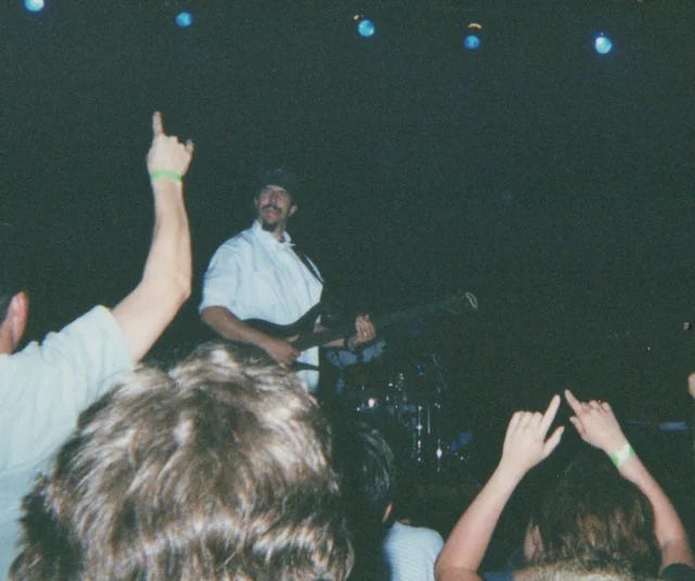 A photo of Les Claypool holding a bass and making a weird face with some fans in the foreground with their hands up.
