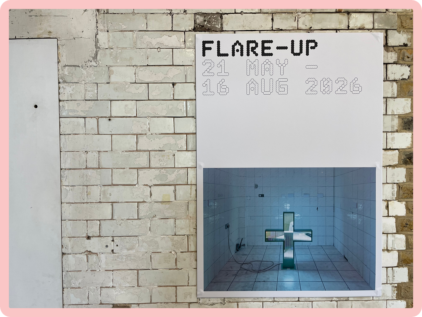 On a white brick wall, a white exhibition poster reads "FLARE-UP, 21 MAY- 16 AUGUST." in a bold, unusual font. Below the title is a photo of one of the exhibition's works: a cross, lit up with an LED, against a white tiled floor and three white tiled walls, giving the impression almost of a bathroom. On a white brick wall, a white exhibition poster reads "FLARE-UP, 21 MAY- 16 AUGUST." in a bold, unusual font. Below the title is a photo of one of the exhibition's works: a cross, lit up with an LED, against a white tiled floor and three white tiled walls, giving the impression almost of a bathroom.