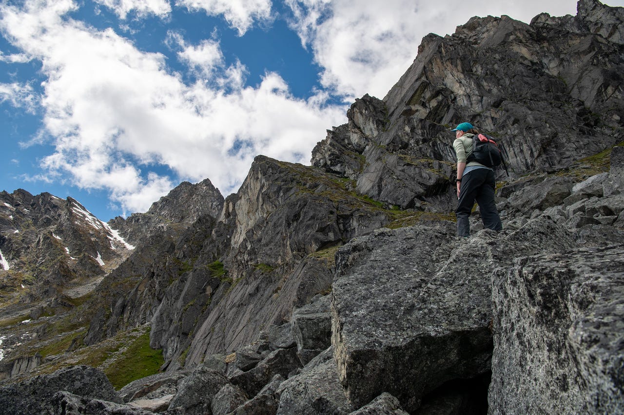 A hiker in a teal cap, pale green hoody, and dark pants climbs away from the camera up a rib of large angular boulders, one hand down for balance. Ahead, tiers of fractured grey cliffs rise toward the skyline, and a snow-holding peak stands at far left under a blue sky with rows of white cloud. A hiker in a teal cap, pale green hoody, and dark pants climbs away from the camera up a rib of large angular boulders, one hand down for balance. Ahead, tiers of fractured grey cliffs rise toward the skyline, and a snow-holding peak stands at far left under a blue sky with rows of white cloud.