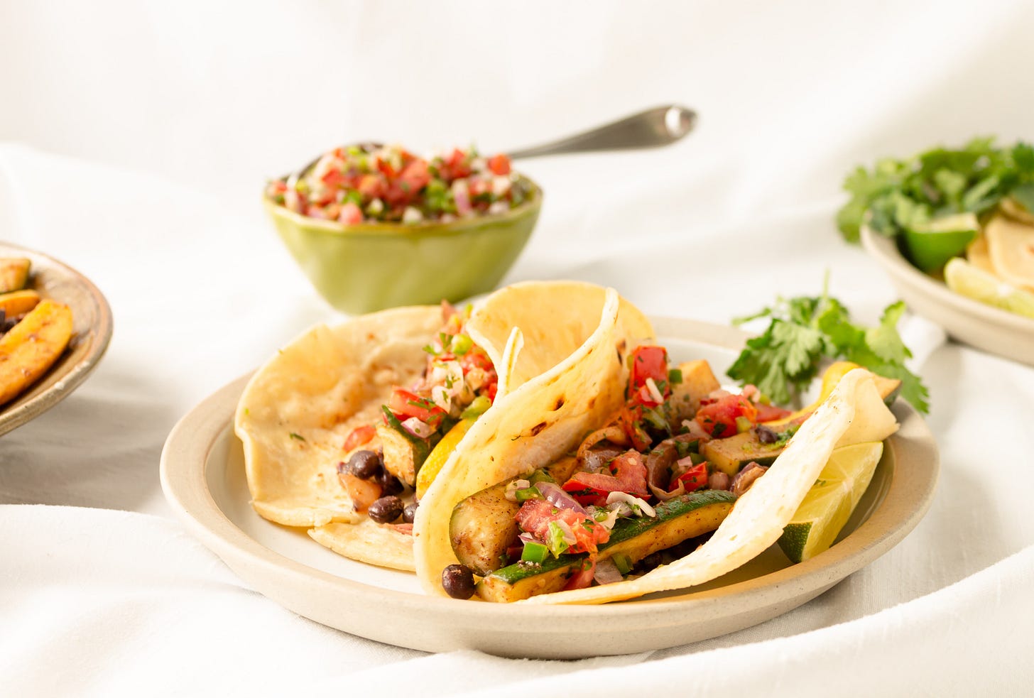 Two gorgeous vegan tacos on cream colored plate. In the background is a delicious looking pico de gallo and other toppings. Two gorgeous vegan tacos on cream colored plate. In the background is a delicious looking pico de gallo and other toppings.