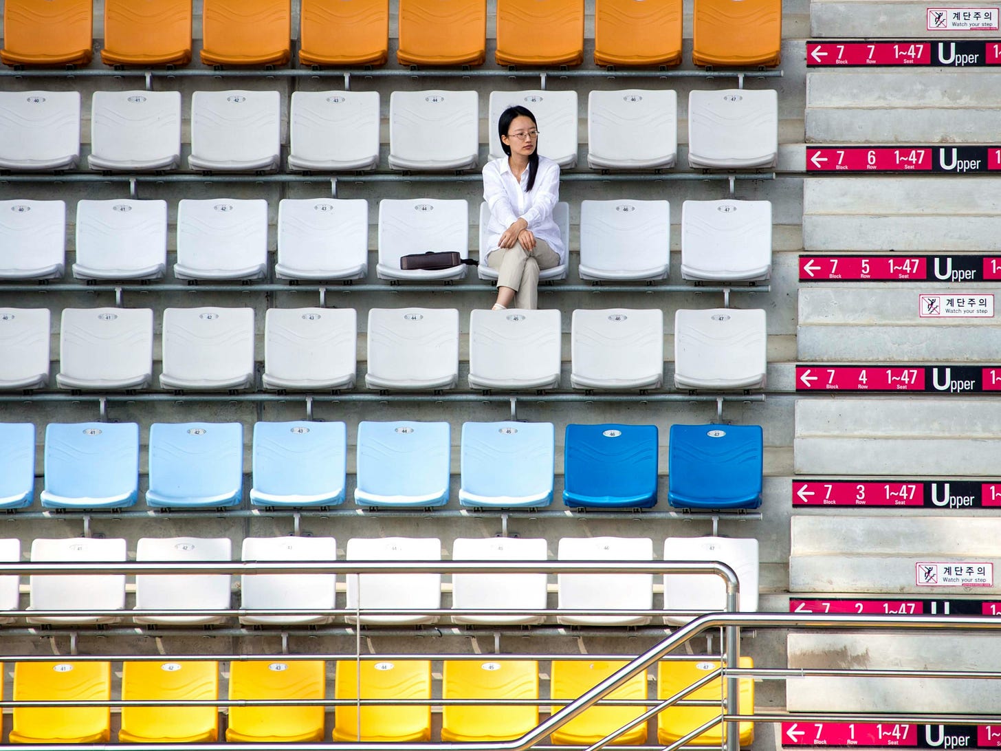 The feedback loop that doesn't close. Woman sitting alone in an empty stadium. The Layer Beneath on Substack.
