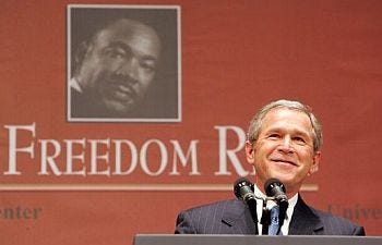 A photo of a smiling George W. Bush standing at a podium, with a photo of Martin Luther King, Jr. behind him. A photo of a smiling George W. Bush standing at a podium, with a photo of Martin Luther King, Jr. behind him.