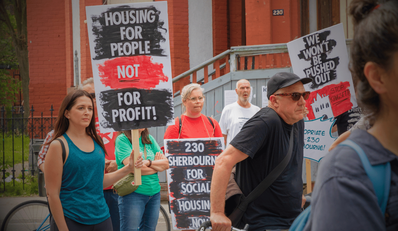 Protesters outside a brick building holding placards reading “Housing for people not for profit” and “We won’t be pushed out.” Protesters outside a brick building holding placards reading “Housing for people not for profit” and “We won’t be pushed out.”